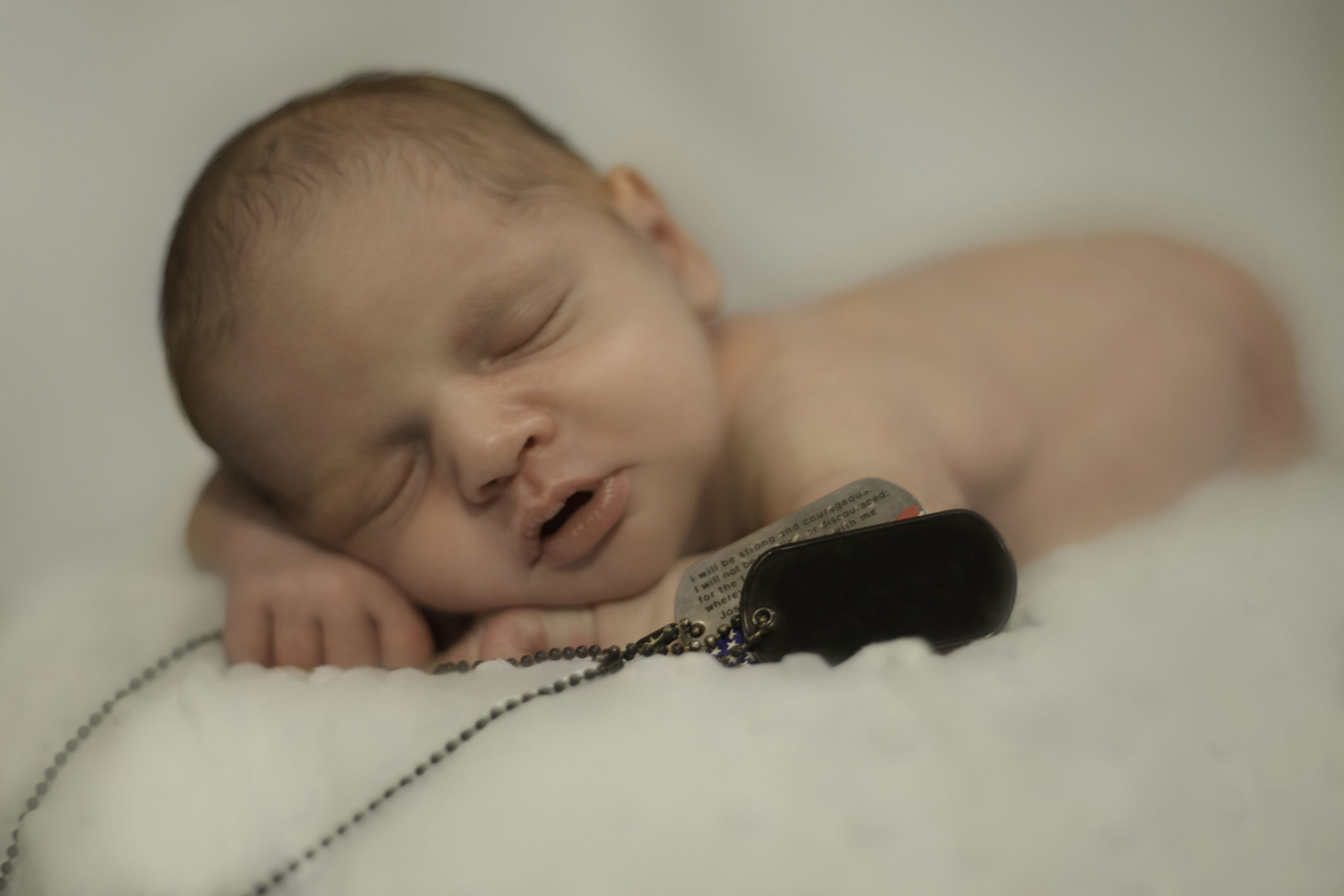 The images shows a newborn baby holding his father's dog tags in remembrance. Image take by Cedar Rapids newborn photographer, Ali Kerr.