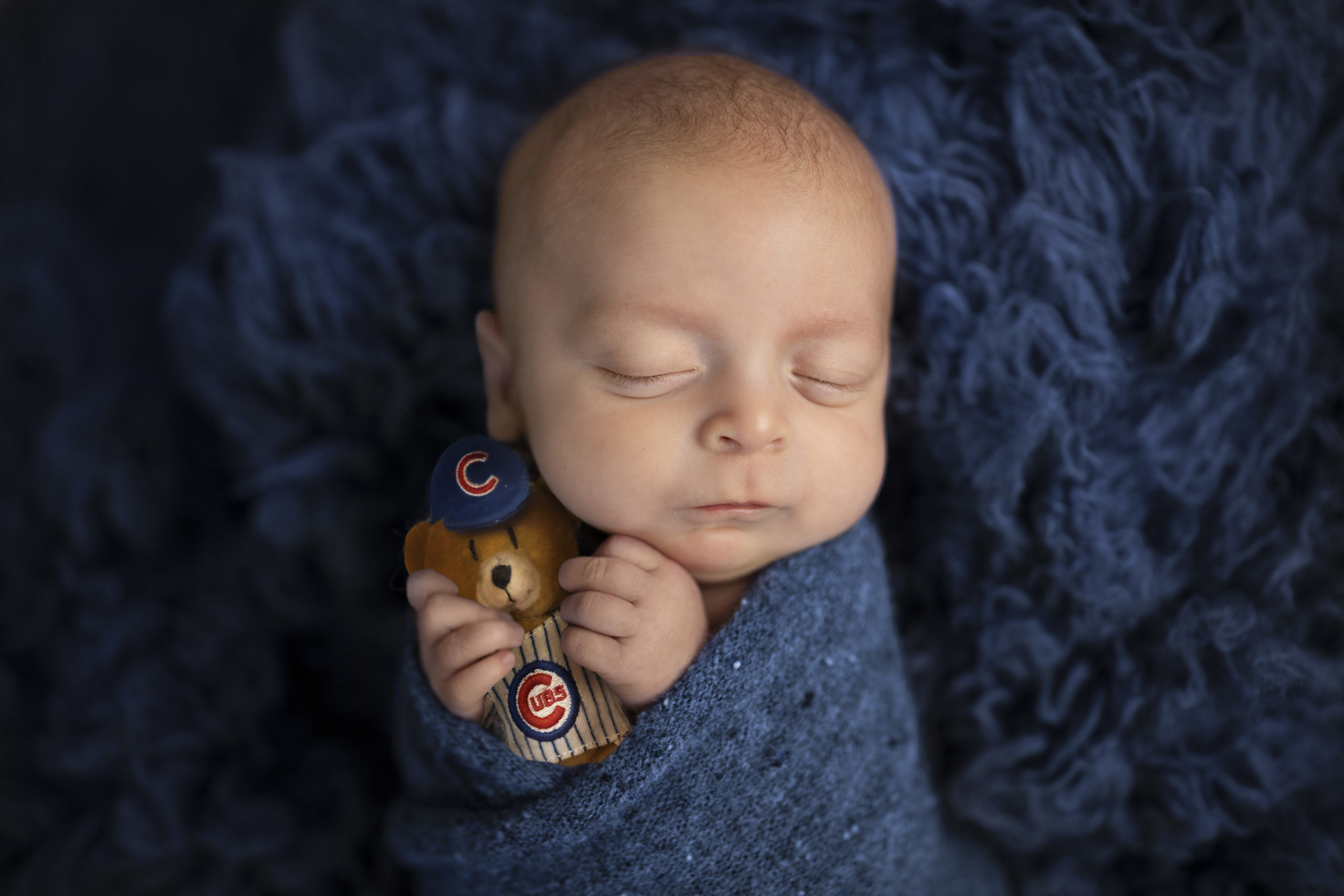 The image shows a newborn baby swaddled holding a Cubs bear. 